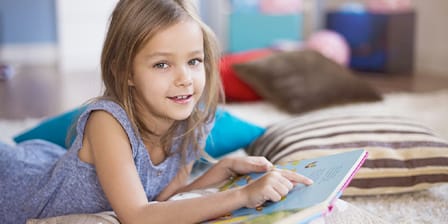 Petite fille allongée sur des coussins en train de pointer du doigt une page de son livre